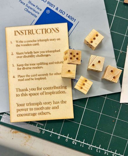 A laser-etched wooden instruction card asking visitors to write a short triumph story, beside six small wooden dice on a cutting mat.